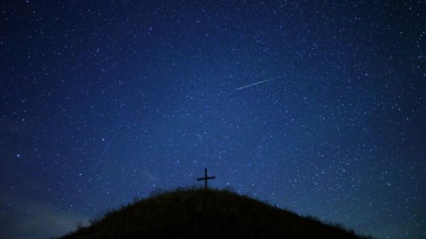 Two meteors streak across a blue night sky, with a hill with a cross on the top in shadow