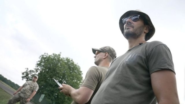 Anton, a drone operator, stands outside in a hat and sunglasses with a man operating a drone behind him